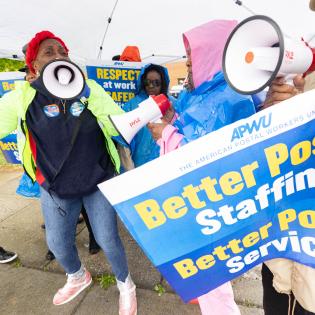 Postal workers rally under a tent in the rain holding rally signs that say better postal staffing, better postal service