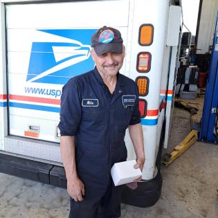 Man in a uniform for maintenance workers wears an APWU sticker on his hat in front of a USPS delivery truck