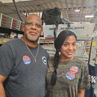 two workers pose in a mail facility with apwu stickers on 