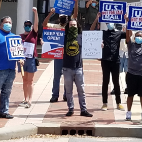 Crowd stands outside a Post Office holding "U.S. Mail Not For Sale" signs. In the center a man in an APWU tshirt holds two signs. one says "keep em open" the other says USMC veterans 4 USPS workers